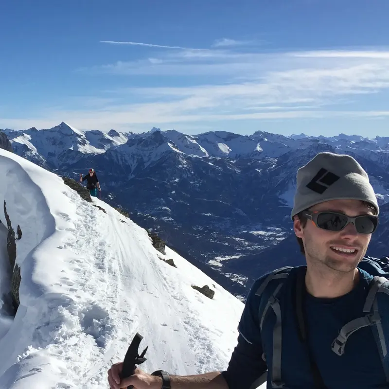 Twee wandelaars lopen over een besneeuwde bergkam onder een strakblauwe hemel; één van hen staat dicht bij de camera en draagt een zonnebril en een muts, terwijl op de achtergrond besneeuwde bergtoppen te zien zijn.